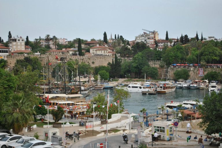 Bustling Antalya harbor with boats, historic buildings, and lush greenery.