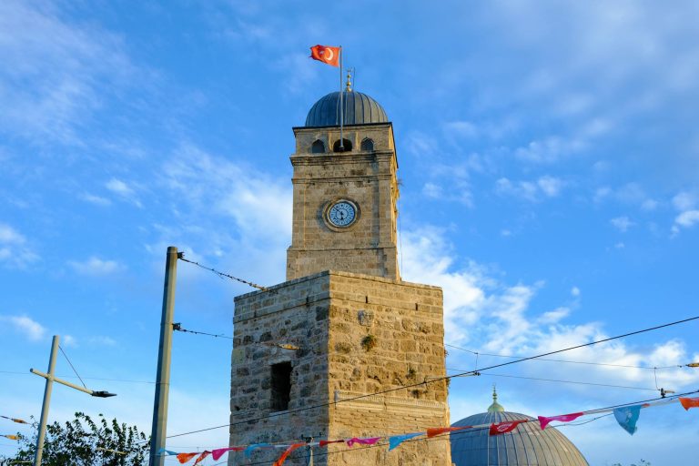 Stone clock tower in Antalya, Türkiye, under a clear blue sky with Turkish flag atop.